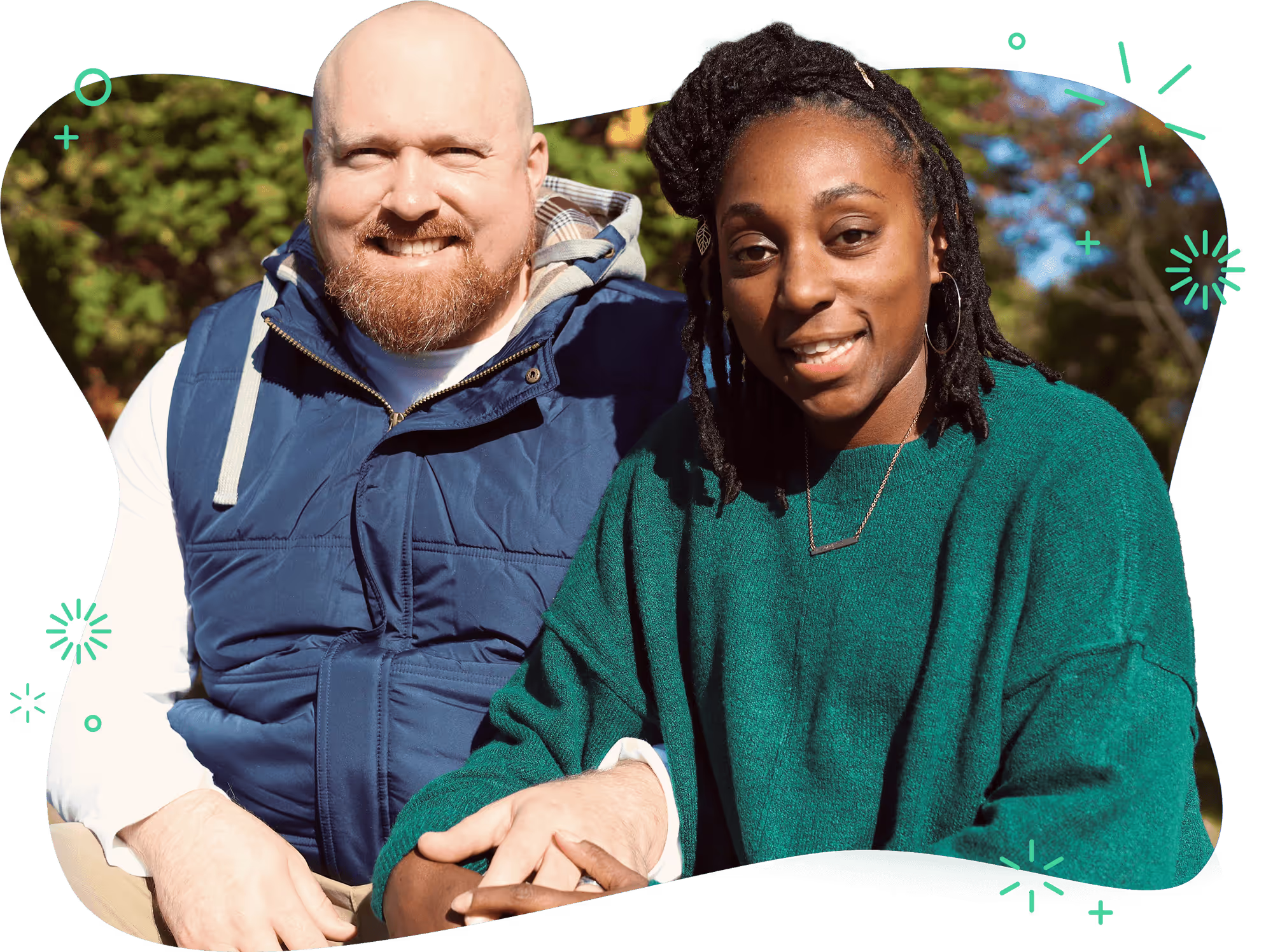 A man in a blue vest sits next to a woman in a green sweater. They're outside holding hands.